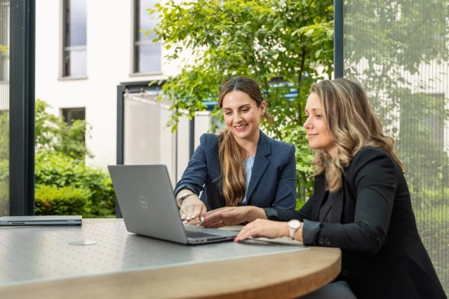 Zwei Frauen am Tisch stehend blicken gemeinsam auf einen Laptop